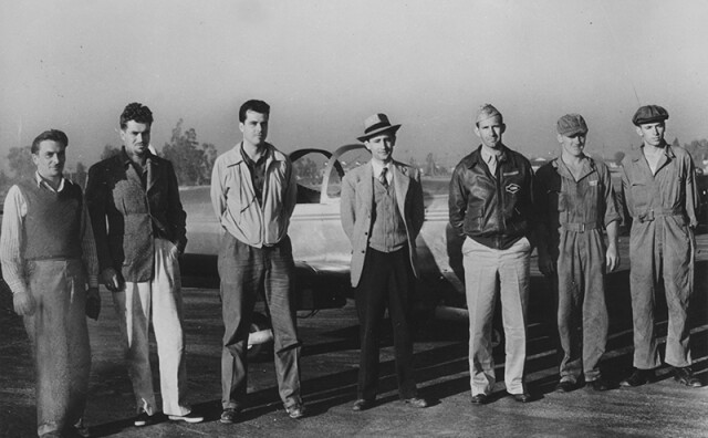 A black and white photo of seven men standing in front of the airplane. Some of them are wearing jackets. A man is in the center wearing a knitted vest with a tie and hat. The man standing next to him on the right is wearing an air force uniform. The two men standing next to him are wearing coveralls and caps.