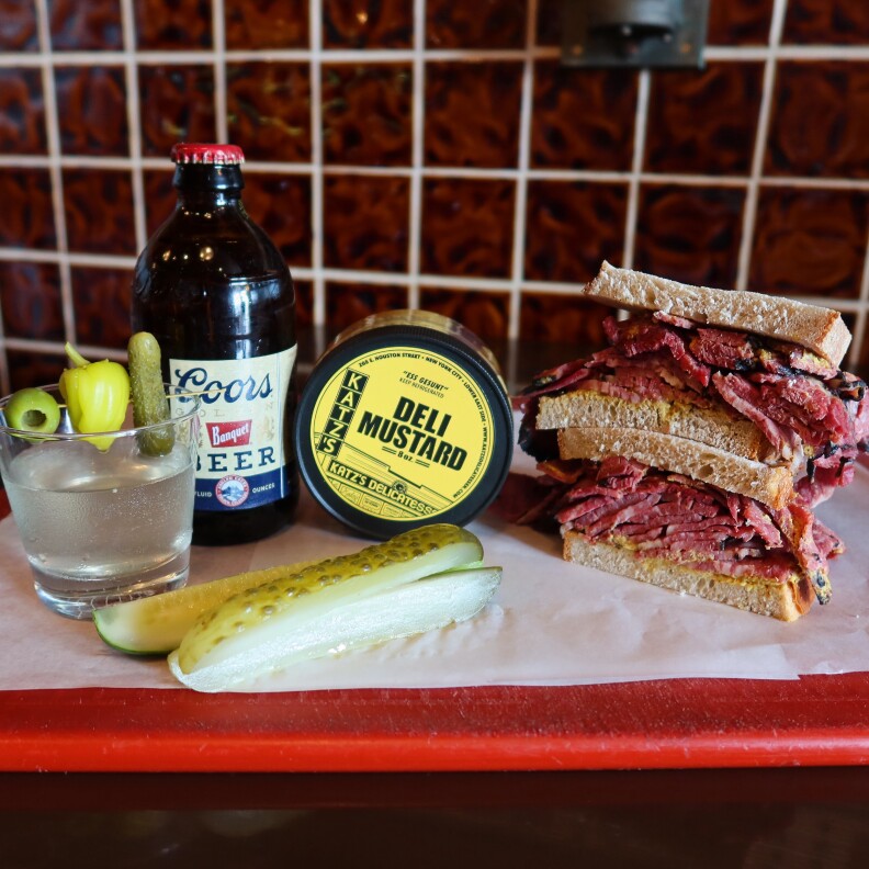 A cocktail with clear liquid is served with an ice cube and garnished with a green olive, yellow pepperoncini, and a small green pickle on a toothpick. Beside it, there's a Coors beer bottle and a canister of Katz's deli mustard. There's also a pastrami sandwich made with light brown bread and two green pickle spears in front. The food is placed on a white surface with a dark red tiled background.