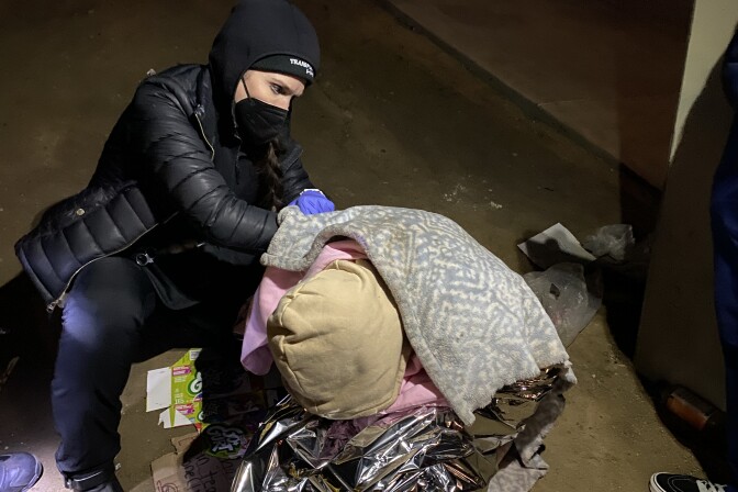 A woman in a black jacket, beenie and wearing a face mask kneels next to a person with a blanket over their back and their hoodie-covered head bowed. 