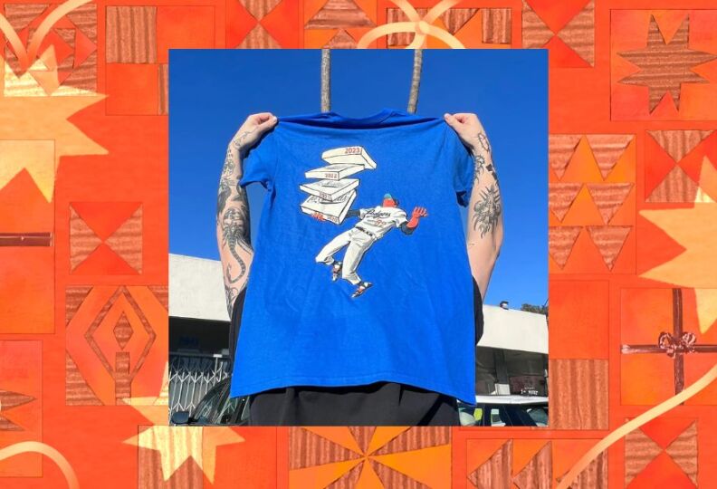 A photograph of a Dodger blue T-shirt being held up over a person's head so that the design is visible: A baseball player in a Dodger's uniform is holding a stack of pizza boxes, as if they all just fell into his outstretched hand in the outfield. The photo is set against a decorative orange-and-gold background, accented with stars, holiday gift boxes and strands of ribbon.