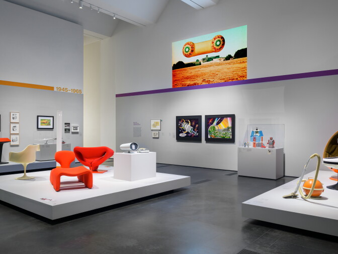 Gallery view of a 1945–1965 section of a design exhibition, with bright orange sculptural chairs and a round orange vacuum cleaner on white platforms, a large projected image of a floating cylindrical structure above a field, and framed artwork and display cases on the walls.