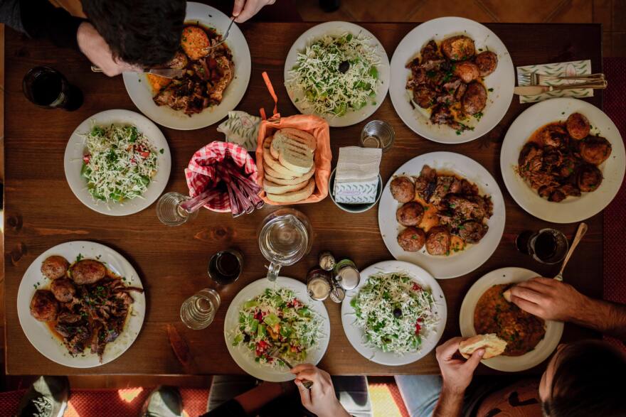 An overhead shot of a dinner table featuring 10 white plates filled with food. Four plates have salad and the rest have sweet potatoes and other dishes. Hands enter the frame with utensils as people eat.
