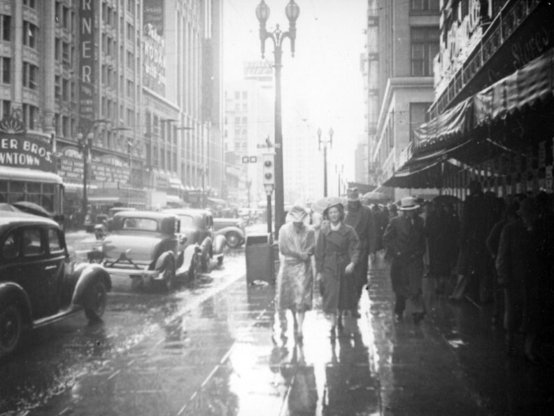 This view looking north on Hill from Seventh Street captures cars driving and people walking in the rain. The movie "Hollywood Hotel" which came out early in 1938 is playing at the Warner Brothers Downtown (previously the Pantages Theater). The Owl Drug Company neon is visible on the right. 