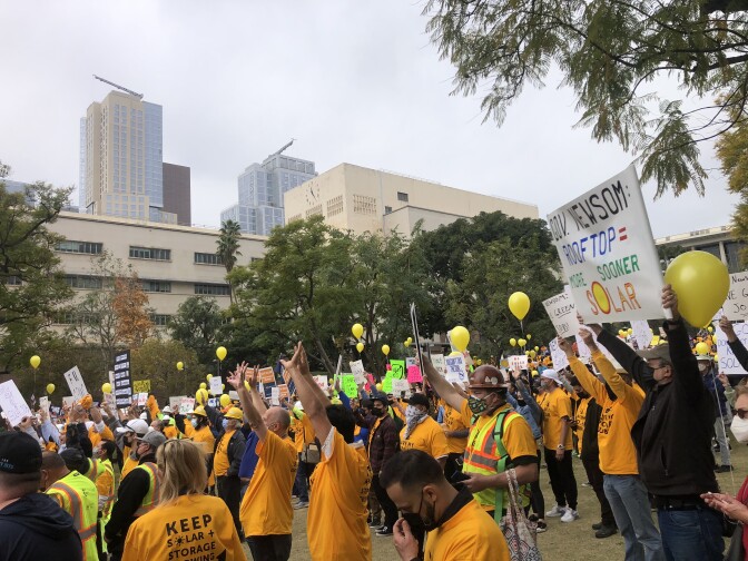 A crowd of people in yellow shirts with signs and yellow balloons cheer with hands up in a grass field in Grand Park in downtown L.A. as buildings loom in the background. 
