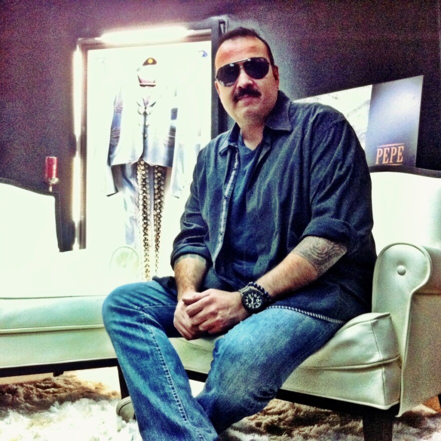 Pepe Aguilar sits on a white chair in a collared shirt with rolled up sleeves over a T-shirt. He wears jeans and a watch and sunglasses. A tattoo is visible on his left arm.