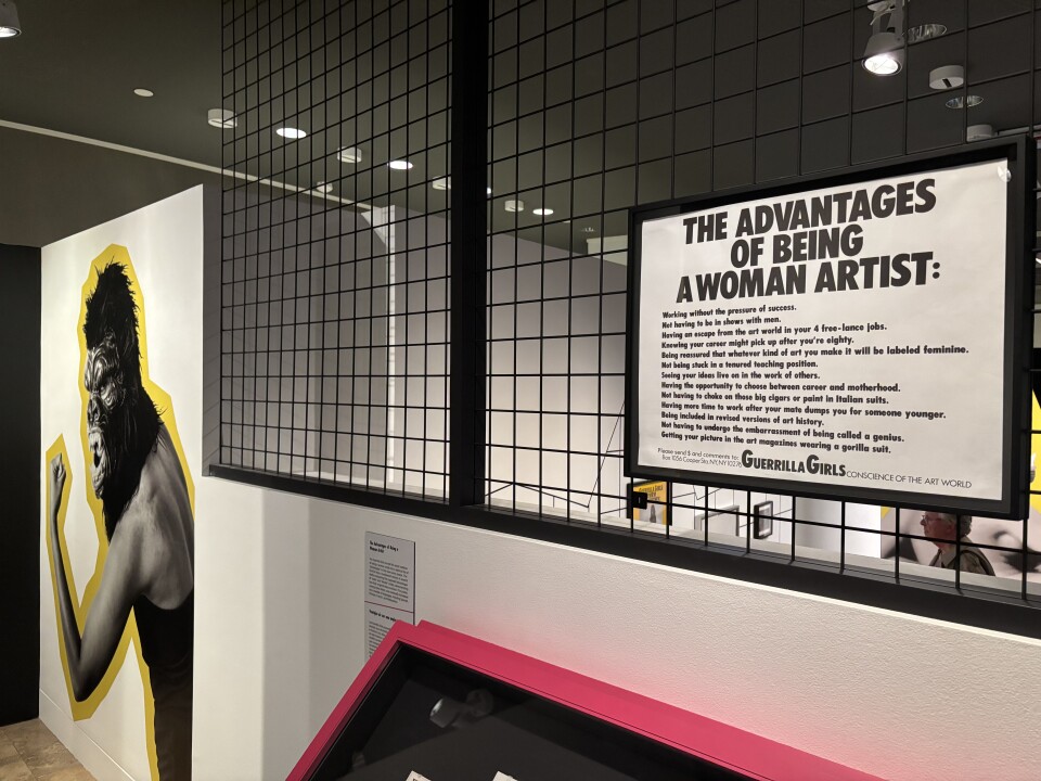 A white half wall with a black metal, cage-like portion on top that reaches to the ceiling. On the metal, a black and white poster titled "The Advantages of Being a Woman Artist" is hung. On the white wall to the left is an enlarged photo of a woman wearing an angry gorilla mask and flexing her bicep.
