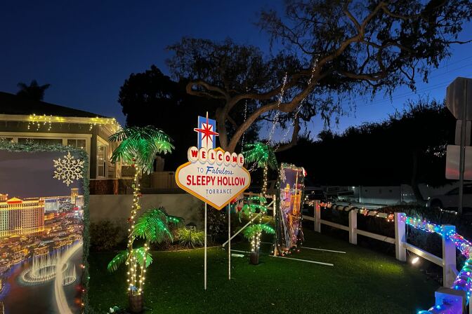 An elaborate holiday light display in the front yard of a home. Lights are wrapped around the white picket fence and fake palm trees, with a large side that reads "Welcome to Fabulous Sleepy Hollow Torrance" sticking out of the green grass in the center. 