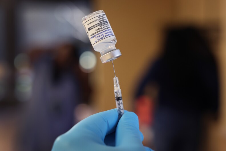 A hand wearing a blue glove inserts a needle into a COVID vaccine glass vial.