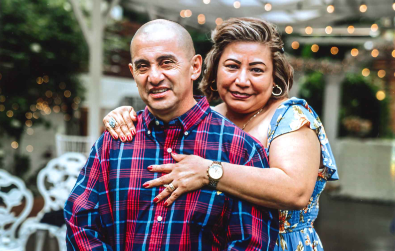 A bald, slightly dark-complected man in a red and blue flannel shirt stands in front of a slightly dark-complected woman wearing a blue dress with colored flowers on it. She is right behind him, with her right hand on his right shoulder and her left hand splayed out in front of his chest, as she displays her wedding ring.