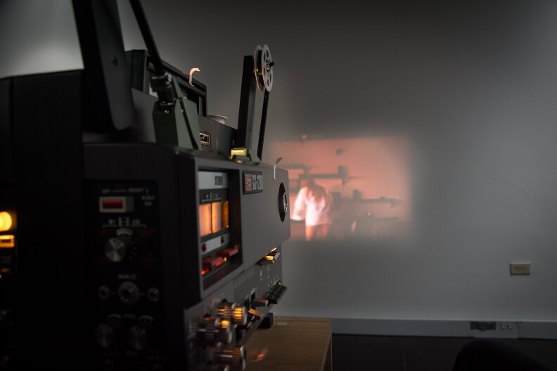 an old movie projector like the kind once used in classrooms projects an image of a person in a white shirt leaning over a desk.