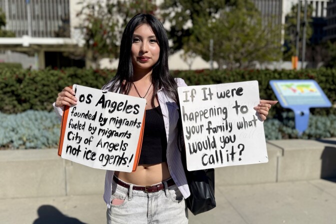 An adolescent girl holds up two signs. One says "Los Angeles, founded by migrants, fueled by migrants, City of Angeles not ICE agents." The other says "If it were happening to your family what would you call it?"