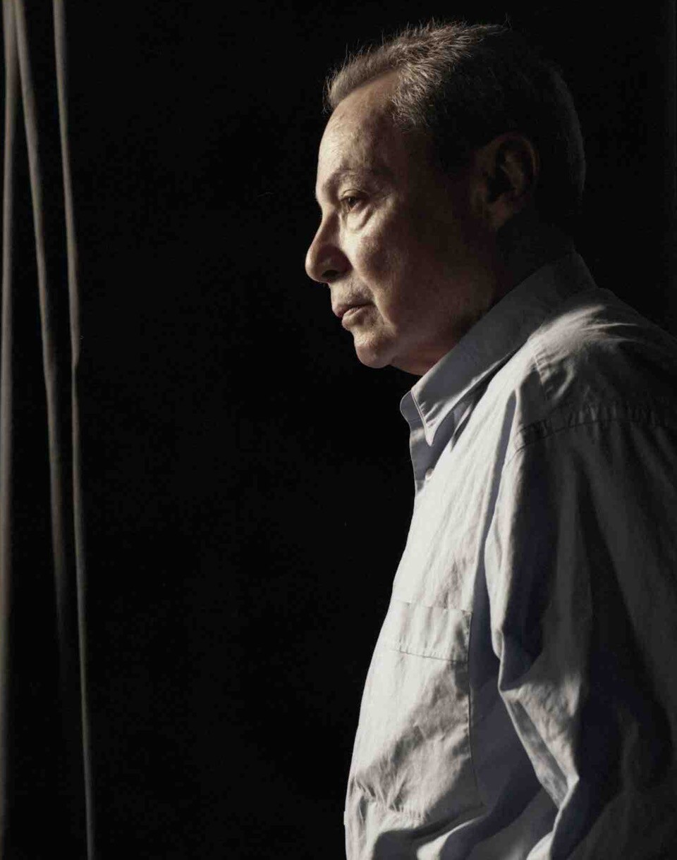 A male presenting person stands and looks to the left. He wears a light colored, button up shirt.