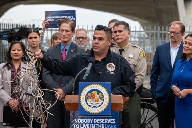 A man stands at a podium speaking into a microphone. In front of the podium, there are two signs with the text: "CALIFORNIA STATE ASSEMBLY" and "NOBODY DESERVES TO LIVE IN THE DARK." In his right hand, the man holds and assortment of tangled wires. Some are yellow, others red and white. There's a group of nine people standing behind him.