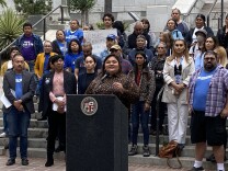 LA City Council member Eunisses Hernandez speaks at a podium that has the seal of Los Angeles on it. There is a crowd of more than a dozen behind her. Some wear blue t-shirts that mark them as supporters of the progressive policy advocacy group LA Forward. 