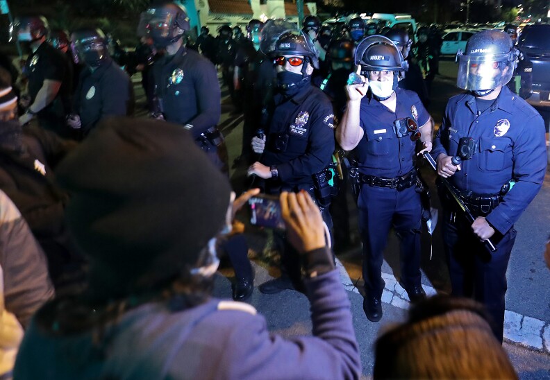 Officers in helmets stand in a line in front of protesters. Once officers appears angry and is gesturing.