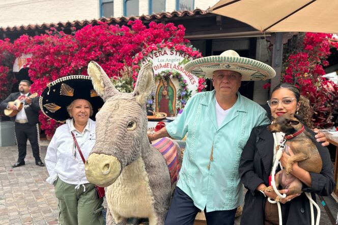 A portrait of three people with medium light skin tones standing near the stuffed donkey. On the sides are two women, who are smiling for the photo. One is holding a dog and the other has a sombrero on. In the middle is Richard, a man with a sombrero on as he puts his hand on the donkey.