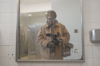 A male presenting man with glasses in a tan jacket looks at himself in a bathroom mirror with a camera pointed towards the mirror.