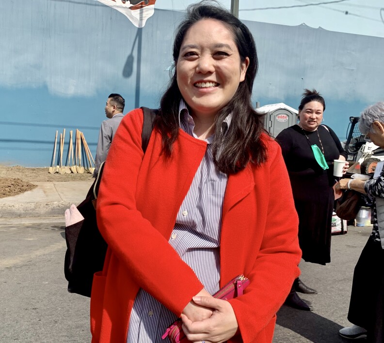 An Asian American woman in her mid-30s stands in the middle of the street with several people around her. She is wearing a striped shirt and red blazer.