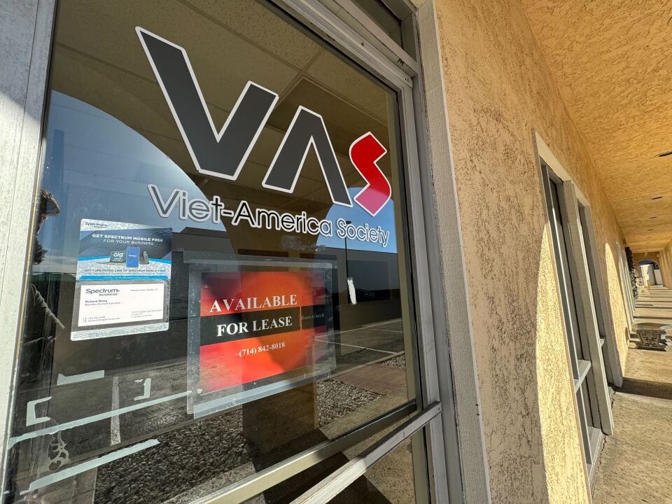 A close-up of a clear glass front door in a long, tan walkway. The door has the text "VAS Viet-America Society" displayed, with an additional sign that says "available for lease" below it.