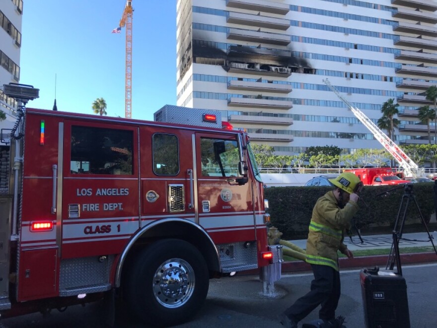 A fire truck sits in the foreground as firefighters work to put out fire and smoke in a high-rise building in the background.