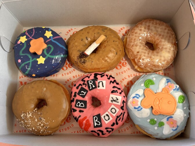 Six doughnuts are lined up in a paper box. The one on the top left has dark blue icing and with yellow stars, the one in the middle has a coffee-brown icing and edible "cigarette" made out of an Italian breadstick. On the bottom, the far right donut is covered in light blue icing and an orange edible fish, with the one next to it covered in pink and black with the words "Burn Book" affixed to the front.