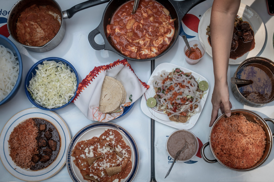 A color photograph taken from above shows several dishes and pans of food arranged on a table, with an arm and hand holding one dish at right.