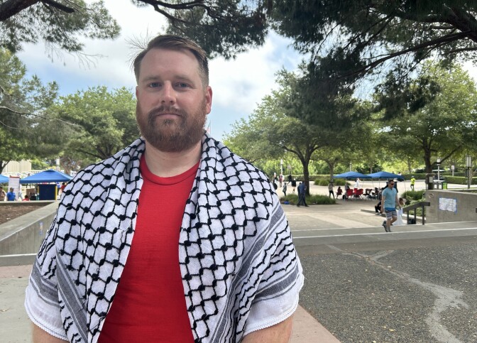 A man with short hair, a beard and mustache, and light skin tone wears a checkered kaffiyeh.