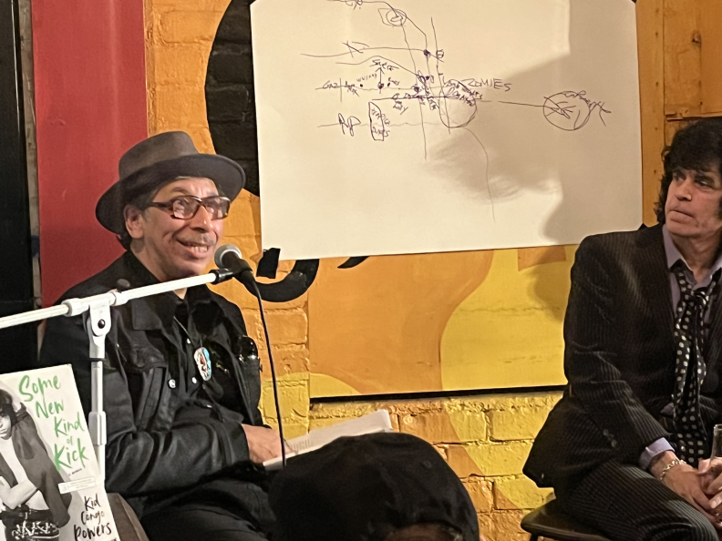 Two men sit in front of a brick wall painted that is red, yellow, and orange. Taped to the wall is a large piece of white paper with a crudely drawn map. The man on the left has medium-dark skin and is wearing a brimmed hat with glasses, a mustache, and all-black clothing. He's speaking into a microphone with a toothy grin on his face. The man to his right has light skin with dark black hair and is wearing a black dotted scarf while listening.