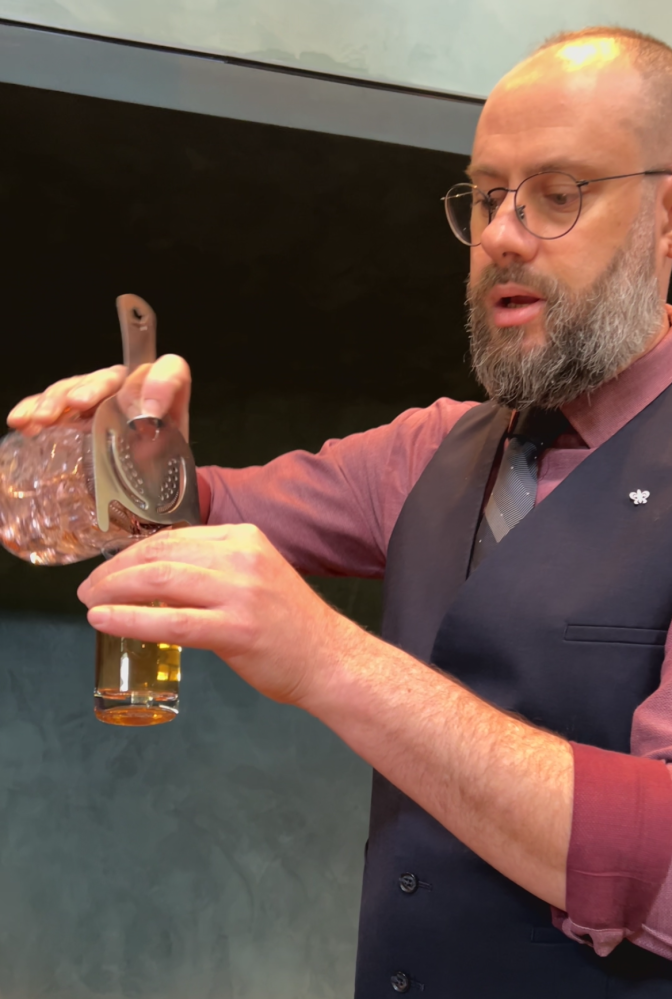 A man with a beard and wearing a shirt, tie and vest pours from a cocktail shaker into a clear glass. 