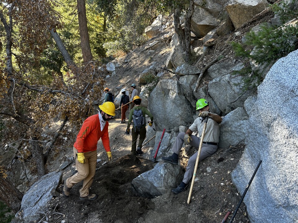 Multiple younger and middle-aged men on a dirt trail with large boulders wearing hardhats and holding McLeod tools do work on a trail. 