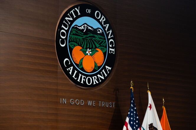 A seal with mountains, rows of farm land, and oranges with the words "County of Orange California" surrounding the scene. The seal hangs on a wooden wall with words inscribed "In God We Trust." At the bottom right of frame there are the ends of three flags.