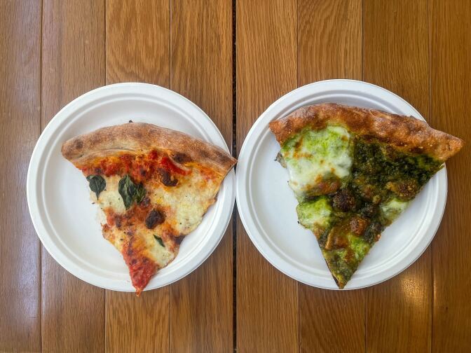 Two paper plates sit side by side, each containing a different slice of pizza. On the left is a pizza slice with red sauce, melted cheese, and cooked basil. On the right is a pizza slice with green sauce and a large amount of melted cheese. Both have light brown crusts at the top.