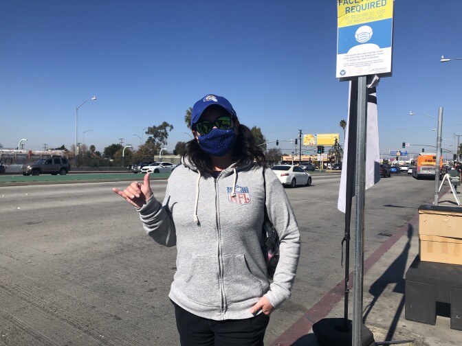 A portrait of Bella Dory. Bella is wearing a blue Rams hat with the Rams logo, an NFL sweatshirt, and a blue and gold mask. She is standing in front of Hawthorne Boulevard, which is busy with buses and cars. Her hand is making the Ram's head hand signal – with her pink and thumb forming the horns. 