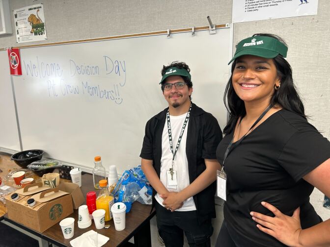 Two people wearing green visors stand in front of a dry erase board