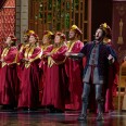 A group of singers in two rows on a stage, with arms crossed in front of them and wearing red robes and hats, both with gold accents. To their left is singer-songwriter Josh Groban in a medeival style outfit with partial knight armor, with his arms outstretched and mouth open in song.