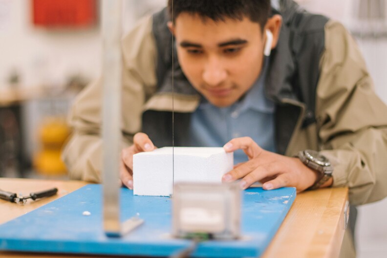 A teenage male student works on a design project in class. He's using tools to neatly slice a block of material while wearing in-ear head phones.