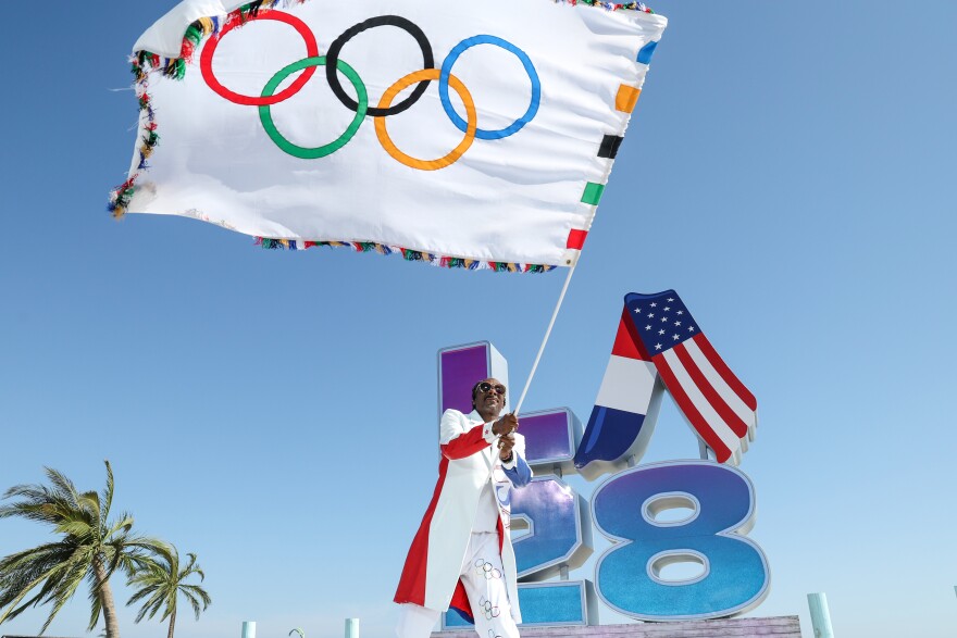 Snoop Dogg waves an Olympic flag. Behind him are palm trees and the logo for the Los Angeles 2028 Olympic games.