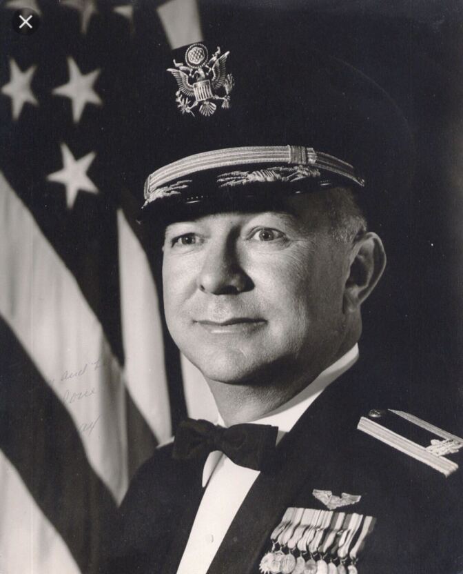 A vintage black and white photo of a man wearing a military uniform in front of an American flag.