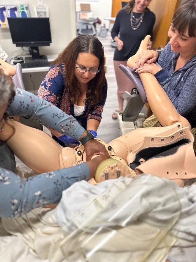 Two women with light skin tone stand at the end of a mannequin in a hospital bed, practicing delivery of a baby.