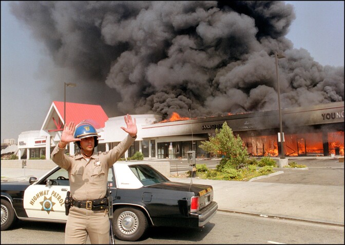 A California Highway patrolman directs raffic around a shopping center engulfed in flames in Los Angeles, 30 April 1992. Riots broke out in Los Angeles, 29 April 1992, after a jury acquitted four police officers accused of beating a black youth, Rodney King, in 1991, hours after the verdict was announced. (Photo credit should read CARLOS SCHIEBECK/AFP/Getty Images)