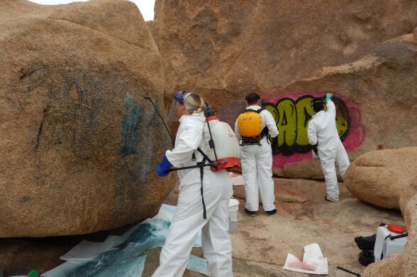 Three people wearing bright-white suits of personal protective equipment are cleaning off spray paint from tan rocks towering over them. Two of the people have containers of liquid on their backs like a backpack, while the third person's arm is outstretched towards the pink and green paint. 