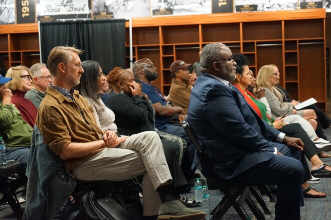 A group of people sit in folding chairs looking straight ahead. A man with a light skin tone, short hair and glasses sits behind a man with a dark skin tone, short hair and glasses.
