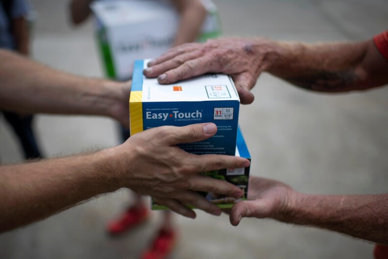 One pair of hairy arms passes a blue box of syringes that say "Easy Touch" in white to another pair of hands that grabs the two boxes.