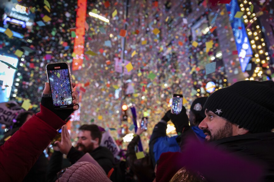 A crowd of people stand underneath falling confetti at night. Some are holding up cell phones, recording the confetti