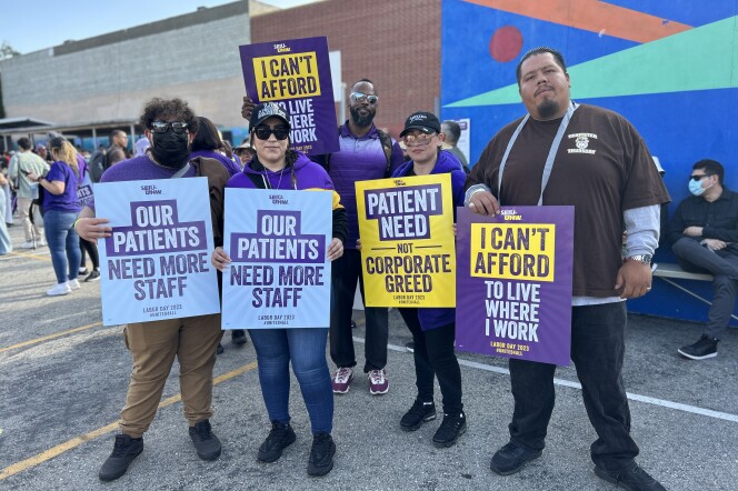 Five people stand holding signs that read "our patients need more staff" "patient need not corporate greed" and "i can't afford to live wehre I work." There are two women with light brown skin, a man with medium brown skin, and man with dark brown skin, and a younger man with light brown skin with curly hair wearing a mask. 