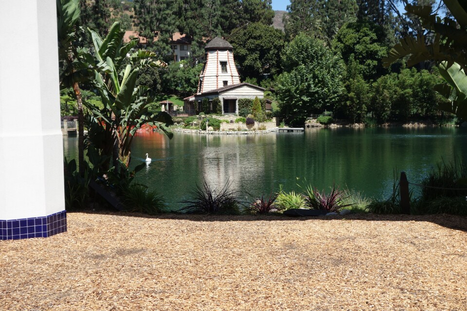 In the foreground, beige sand and a wite pillar with blue tiles at the base. In the background, a windmill. You can see a swan swimming on the lake.