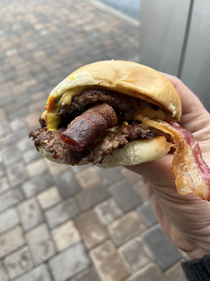A hand with a light skin tone is holding a cheeseburger that features two large pieces of bacon protruding between the burger patties with a small amount of melted yellow American cheese.