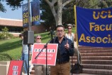 A person with with short hair and reading glasses speaks while gesturing at the crowd. Behind him, people wearing sunglasses hold up a large blue sign that reads: "UCLA Faculty Association" in gold letters. They are on a college campus, with brick steps and grassy hills.
