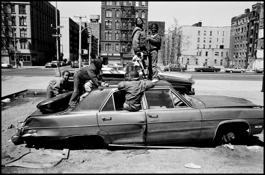 An abandoned sedan is parked on its rims as children climb on and in it.