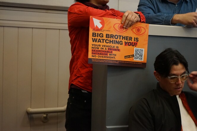 A person with a medium skin tone wearing a red long sleeved shirt leans on a wall holding an orange sign that reads "BIG BROTHER IS WATCHING YOU! Your vehicle is now in a private, searchable database with no oversight."